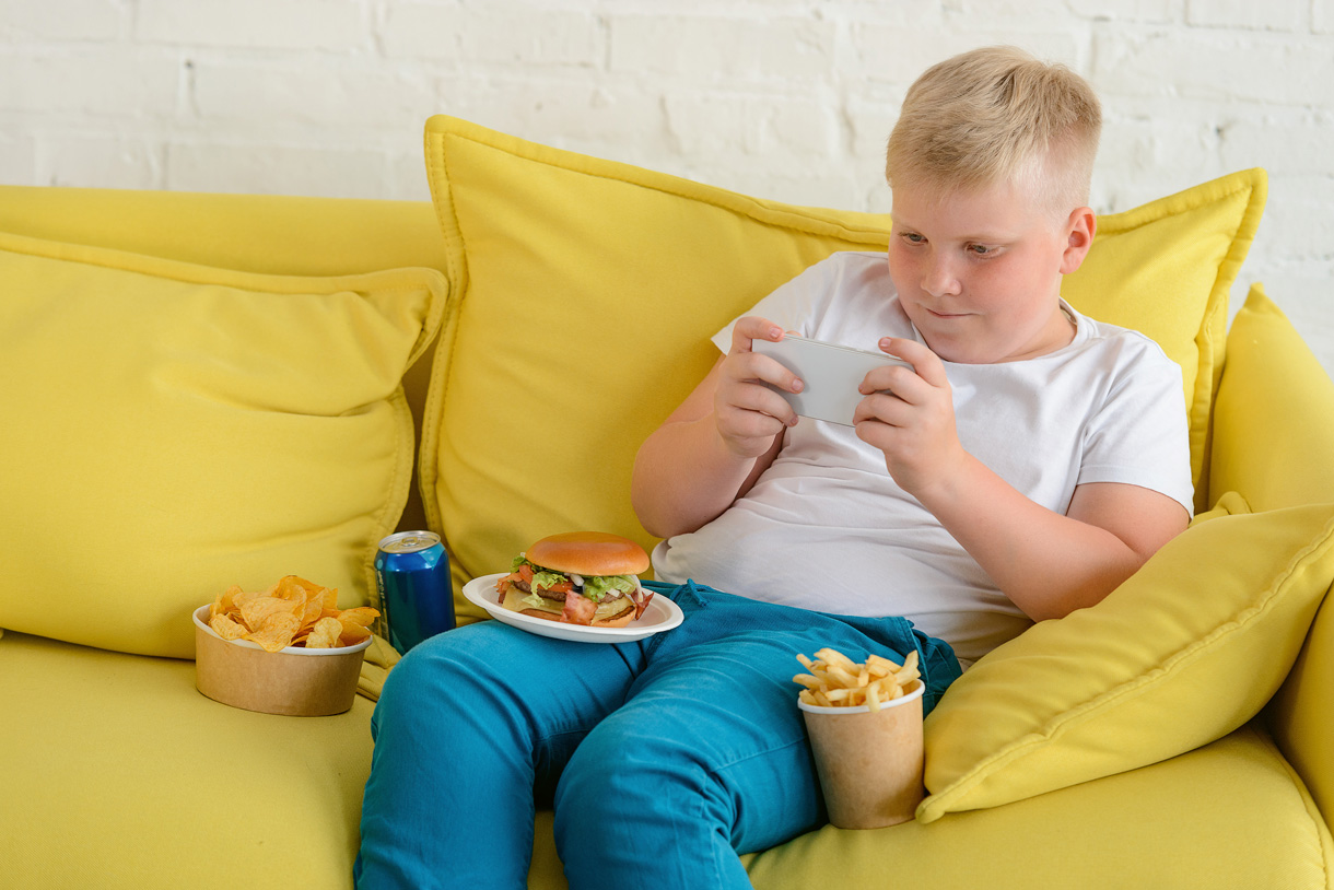 Ein übergewichtiger Junge sitzt mit dem Handy auf dem Sofa. Um ihn herum steht ein Süssgetränk und fast Food.