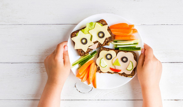 Zwei Vollkornbrotscheiben mit Käse-Sternen, Oliven-Augen und sonstigen frischen Zutaten. So belegt, dass einem zwei freundliche Gsichter anlachen.