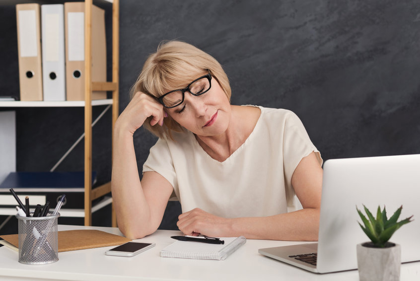Eine Frau sitzt müde am Schreibtisch vor ihrem Laptop. Sie stützt den müden Kopf mit der rechten Hand und ruht mit geschlossen Augen kurz aus.
