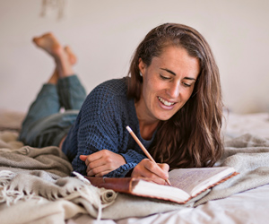 Eine Frau liegt liegt auf dem Bauch auf einem Bett und schreibt gerade gelassen in einem Buch.