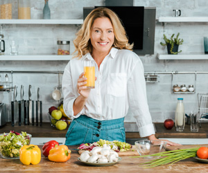 Eine reife Frau steht in der Küche und vor ihr liegen diverse frische Lebensmittel. Sie hält ein Glas Fruchtsaft und lacht zufrieden in die Kamera.