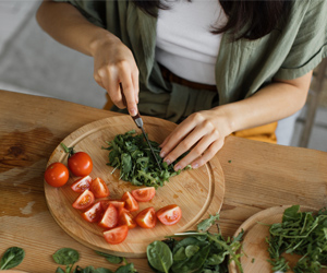 Man sieht aus der Vogelperspektive eine Frau mediterranes Gemüse und Kräuter schneiden. Tomaten, Rucola, Kresse und ihre Hände sind zu sehen.