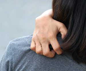 Man sieht die linke Schulter einer Frau mit langen dunklen Haaren. Sie kratzt sich mit der Hand den Rücken.