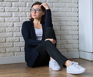 Eine reife Frau sitzt, an eine Wand gelehnt, auf dem Fussboden und sieht nachdenklich in die Ferne.