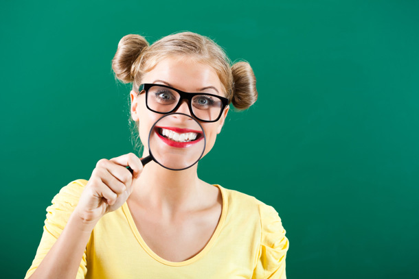 Eine junge Frau mit Brille hält sich vor den Mund eine Lupe. Ihr Mund bzw. sie zeigt die Zähne, werden durch die Lupe etwas vergrössert.