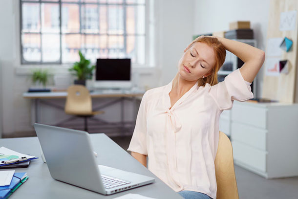 Eine junge Frau sitzt im Büro vor ihrem Laptop und versucht Ihren Nacken zu entspannen. Sie dehnt den Kopf nach links und hält ihn mit dem linken Arm fest.