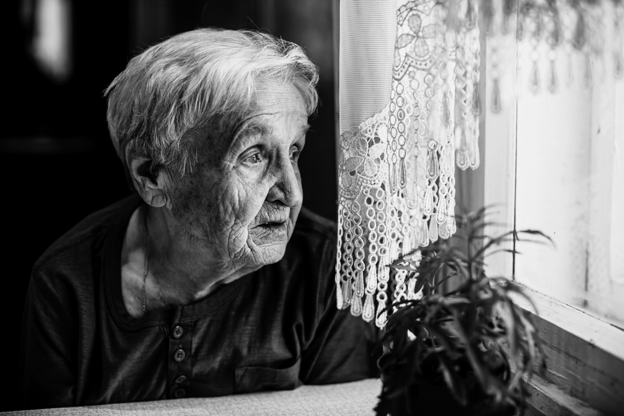 Schwarz-Weiss-Foto: Eine alte Dame mit kurzem Harr schaut aus dem Wohnungsfenster in die Ferne.